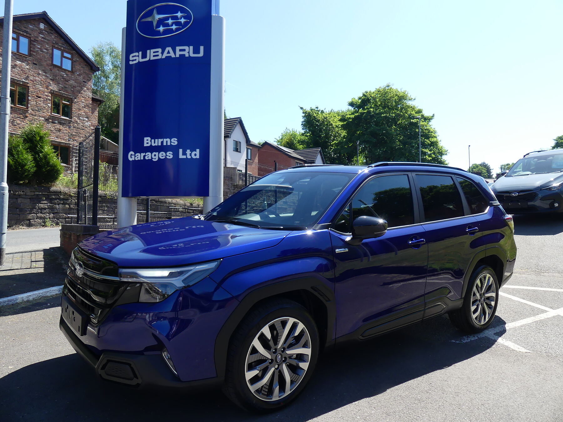 A new Subaru Forester in Medium Sapphire Blue beside the Burns Garages Subaru sign on the forecourt in Congleton
